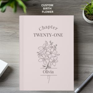 May include: A light pink hardcover book featuring the text "Chapter TWENTY-ONE" and the name "Olivia" printed in dark gray. Below the text is a line-art illustration of a bouquet of flowers. The cover also includes the text "CUSTOM BIRTH FLOWER".