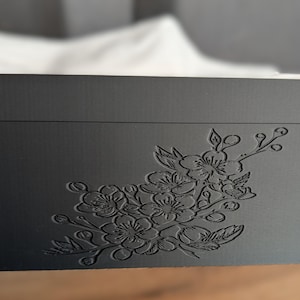 Peut inclure: Boîte à mouchoirs noire de forme rectangulaire, ornée d'un motif floral gravé. Le coffret est en matériau mat et possède un couvercle. Des mouchoirs blancs sont visibles à l'intérieur.