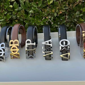 May include: A collection of leather belts with metallic lettered belt buckles. The belts are black and brown, with silver, gold, and bronze-colored buckles spelling out Greek letters and names. The belts are displayed on a white tray.
