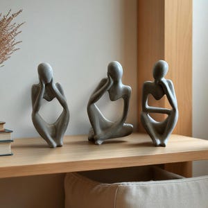 Pode incluir: Três esculturas cinzentas de forma humana abstrata em poses sentadas. Cada figura apresenta uma forma lisa com centros ocos, representando indivíduos em posições contemplativas com as mãos perto da cabeça ou do queixo.