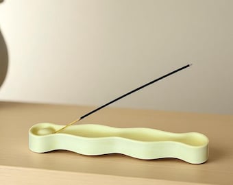 May include: A pale yellow, elongated, wavy-shaped ceramic incense holder with a circular indentation at one end. A single black incense stick with a wooden base is inserted into the holder, angled diagonally upward from the circular depression.