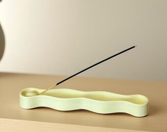 May include: A pale yellow, elongated, wavy-shaped ceramic incense holder with a circular indentation at one end. A single black incense stick with a wooden base is inserted into the holder, angled diagonally upward from the circular depression.