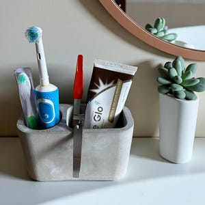 Puede incluir: Un portacepillos de dientes de hormigón gris con un divisor metálico contiene un cepillo de dientes eléctrico azul, un objeto rojo y un tubo de pasta de dientes. A la derecha hay una pequeña maceta blanca con una suculenta. Un espejo redondo en el fondo.