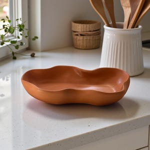 May include: A terracotta-coloured ceramic bowl with an irregular, organic, wavy rim shape. The bowl has a smooth, matte finish and a shallow, wide basin, resting on a flat surface. The design features soft, undulating curves along the entire edge.