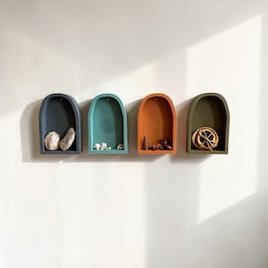Puede incluir: Cuatro estantes de pared en forma de arco en colores carbón, azul cerceta, terracota y verde oliva. Cada estante contiene pequeños objetos decorativos, incluyendo piedras claras, conchas blancas, un cono de incienso marrón y una rodaja de cítrico seca.