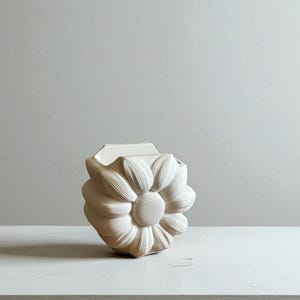 May include: A cream-colored, ceramic vase with a daisy-like design. The vase has a square top and a rounded, flower-shaped body with detailed petal impressions. The center of the flower is textured, and the vase sits on a white surface.