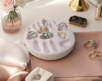 Peut inclure: Un plateau organisateur de bijoux rond, de couleur lavande pâle, doté de multiples fentes encastrées pour ranger des bagues. Le plateau est présenté avec diverses bagues métalliques placées dans les fentes, ainsi que d'autres bagues sur un tissu rose.
