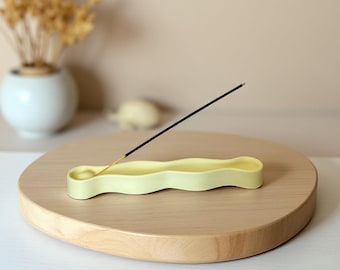 May include: A pale yellow, elongated, wavy-shaped incense holder with a circular indentation at one end. A single black incense stick with a glowing orange tip is inserted into the holder, resting at an angle against the smooth, matte surface.