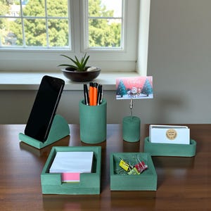 May include: A set of green concrete desk tidies. The collection includes a phone stand, pen pot, card holder, business card holder, a square tray for paper, and a small tray for stationery. A Christmas card is in the card holder.