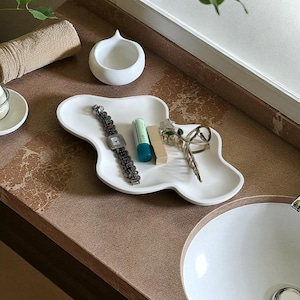 May include: An irregular white tray holds a silver-toned watch with a dark link band, a small green and white tube, a rectangular tan object, and a silver-toned hair clip. Nearby are a small white bowl, a white dish with cotton balls, and a tan towel.