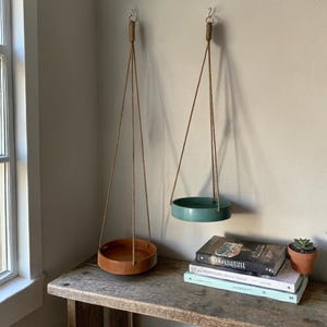 May include: Two hanging planters with brown leather-like bases and teal-coloured bases suspended by rope. The planters are hanging from hooks on a neutral-coloured wall. The planters are empty, ready for plants.