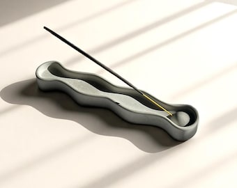 May include: A light gray, wavy, elongated incense holder made of a smooth material. The holder features a central channel with a circular indentation at one end, where a thin black incense stick with a yellow core is positioned at an angle.