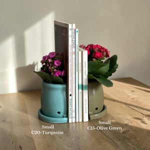 May include: Two small ceramic pots, one turquoise and one olive green, each holding a flowering plant. Books stand between the pots. The text on the pots reads "Small C20-Turquoise" and "Small C15-Olive Green".