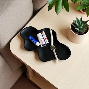 Op de afbeelding: Een zwart, organisch gevormd dienblad bevat verschillende items. Een blauwe tube, een klein flesje nagellak en twee lippenbalsems zijn op het dienblad geplaatst. Een vierkant horloge met een crèmekleurige band ligt op het dienblad. Een kleine vetplant in een pot staat op de achtergrond.