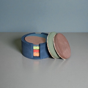 May include: A set of round coasters in a blue holder. The coasters are in shades of brown, green, and orange. The holder is a dark blue with a circular design. The coasters are made of a solid material.