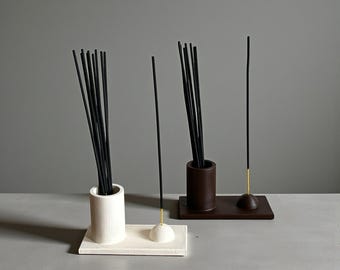 May include: Two incense holders, one white and one dark brown, each featuring a cylindrical vessel and a small dome-shaped base. Both holders contain bundles of thin black incense sticks, with one single stick placed upright in each dome-shaped base.