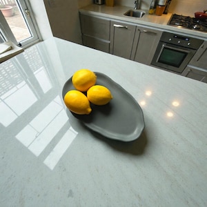 Peut inclure: Trois citrons jaunes vifs sont disposés sur un plat en céramique gris de forme ovale. Le plat est posé sur un plan de travail de cuisine de couleur claire. Les citrons attirent l'attention.