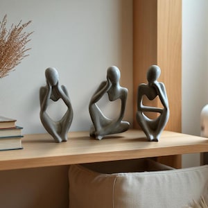 Pode incluir: Três esculturas cinzentas com forma humana abstrata em poses sentadas, cada uma com um acabamento liso e um design de torso oco. As figuras estão posicionadas lado a lado, com cada escultura exibindo uma postura contemplativa diferente.