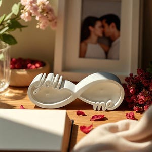 Peut inclure: Une sculpture blanche en forme de symbole infini avec les mots "you" et "me" sur une surface en bois. La sculpture est entourée de pétales de roses rouges, d'une photo encadrée d'un couple s'embrassant et de compositions florales.