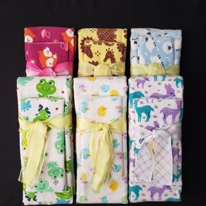 May include: A collection of baby washcloth sets, each wrapped with a ribbon. The sets feature various animal prints, including foxes, giraffes, frogs, ducks, deer, and bears. The washcloths are in shades of pink, yellow, blue, and white.