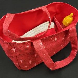 Peut inclure: Sac fourre-tout en tissu rouge avec un motif d'étoiles blanches et roses. Le sac est ouvert et rempli de couches et d'un biberon jaune.