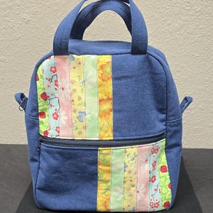 May include: A blue denim lunch bag with a colorful floral patchwork design on the front. The patchwork includes vertical stripes of various floral patterns and colors, including red ladybugs, yellow sunflowers, and pink flowers. The bag has a zipper closure and a top handle.