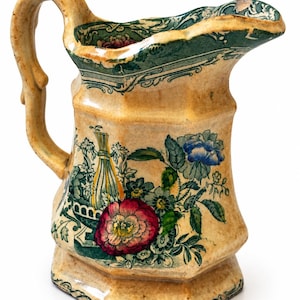 May include: A cream-colored ceramic pitcher with a hexagonal body and a curved handle. The surface features a transfer-printed design of green foliage, a large red flower, a blue flower, and a tall yellow wheat sheaf, with a green border at the rim.