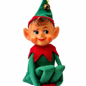 May include: A vintage-style elf figurine wearing a green felt suit with a red collar and red shoes. The elf has a painted face with blue eyes, rosy cheeks, and brown hair. A green pointed hat with a small metal bell and a yellow string is attached.