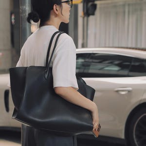 Może przedstawiać: Duża, czarna torba typu tote z długimi uchwytami. Torba jest trzymana przez osobę ubraną w biały t-shirt i ciemne spodnie. Tło obejmuje biały samoch&oacute;d i scenę uliczną.