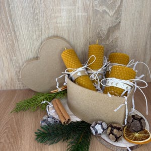 Peut inclure: Trois bougies en cire d'abeille à texture alvéolée jaune, entourées de ficelle blanche à motifs de petits cœurs, disposées dans une boîte en carton en forme de cœur avec des bâtons de cannelle, des aiguilles de pin, des tranches d'orange séchées et des pommes de pin.