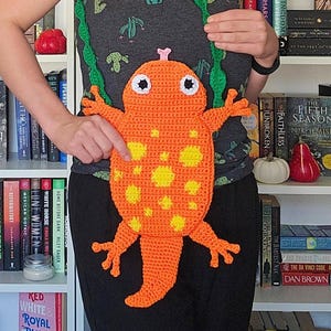 Könnte beinhalten: Eine orangefarbene, gehäkelte Frosch-Tasche mit gelben Punkten und weißen Augen. Die Tasche hat einen grünen Riemen und wird über einem grauen Tanktop mit Kaktus-Design getragen. Handgefertigt und verspielt.