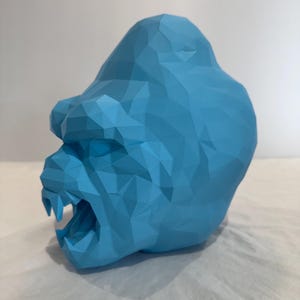 May include: A light blue, geometric gorilla head sculpture. The sculpture has a polygonal design with an open mouth, revealing teeth. The gorilla is set against a white background.