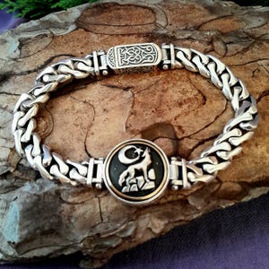May include: Silver chain bracelet with a decorative clasp and a circular centerpiece. The centerpiece features a wolf and crescent moon design. The bracelet is displayed on a textured, brown surface.