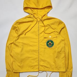 May include: A vibrant yellow hooded jacket with a full-length zip and a small chest pocket. The jacket has a patch with the words "Troop Committee" and a Boy Scout emblem. It features a drawstring at the hem and long sleeves.