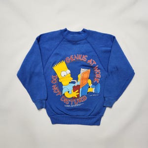 Könnte beinhalten: Blaues Sweatshirt mit Bart Simpson, der ein Buch liest und trinkt. Die Grafik enthält den Text "GENIUS AT WORK" und "DO NOT DISTURB". Das Sweatshirt hat einen Rundhalsausschnitt.