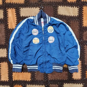 Può includere: Giacca da baseball blu vintage con strisce bianche sulle maniche e sul colletto. La giacca presenta toppe per i Phillies, gli Yankees e altre squadre. Ha una chiusura con cerniera e polsini e orlo a coste.