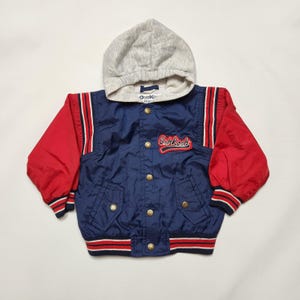 May include: A navy blue jacket with red sleeves, a light grey hood, and a front button closure. The jacket features the red and white embroidered text OshKosh on the chest, two front pockets, and striped ribbed cuffs and hem in red and white.
