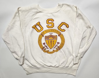 May include: Cream-colored sweatshirt featuring the letters "USC" in gold and maroon. The sweatshirt displays the University of Southern California emblem, a shield with a sunburst, torch, and roses.