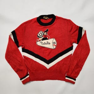 May include: Red sweater with black and white accents. Features a graphic of a person on a log with the name "Natalie" and the number "85". The sweater has a crew neck and striped cuffs.