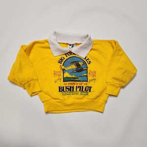 May include: A bright yellow long-sleeve pullover shirt with a white collar. The front features a graphic of a yellow floatplane over water with the text "BIG FOOT LAKES," "River Trips," "Pack Trips," "FLY NORTH OF 51° LAT," and "BUSH PILOT REGISTERED GUIDE."