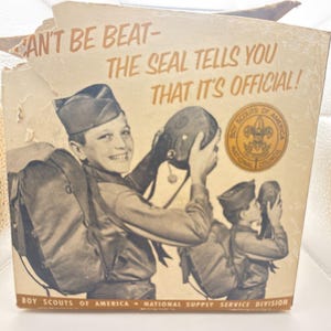 Op de afbeelding: Vintage Boy Scouts of America doos met illustraties van scouts. De doos bevat de tekst "CAN'T BE BEAT - THE SEAL TELLS YOU THAT IT'S OFFICIAL!" en het logo van de Boy Scouts of America. De doos is bruin en beige.