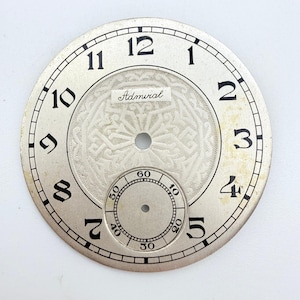 May include: A circular silver-toned watch dial featuring black Arabic numerals from 1 to 12. The center displays an intricate guilloché pattern with the word Admiral printed in black. A smaller sub-dial at the bottom tracks seconds from 10 to 60.