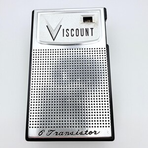 Puede incluir: Una radio portátil rectangular Viscount con marco de plástico negro y panel frontal de metal plateado con patrón de rejilla. La parte superior muestra la marca Viscount y la parte inferior tiene la inscripción 6 Transistor.