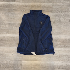 May include: Navy blue fleece jacket with a full zipper and two side pockets. The jacket features a high collar and a small embroidered design on the chest. A tag is attached to the bottom of the jacket.