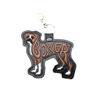 May include: A Boxer dog-shaped keychain with the word "BOXER" embroidered in brown. The dog has brown and white markings, a black outline, and a silver-colored clasp.