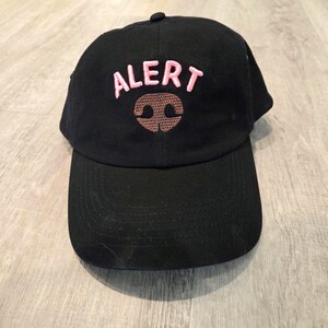 以下が含まれることがあります： ピンク色の刺繍で「ALERT」の文字と茶色の犬の鼻のデザインが施された黒い野球帽。 つばがカーブしており、上部にボタンが付いています。
