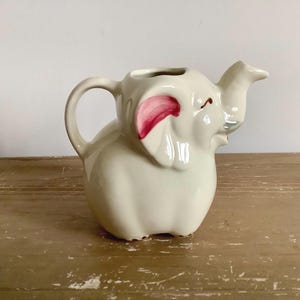 Peut inclure: Pichet en céramique crème en forme d'éléphant, avec une anse et un bec verseur. L'éléphant a des accents roses sur les oreilles et une petite trompe incurvée. Le pichet est posé sur une surface en bois.