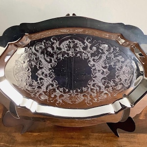 May include: An oval silver-toned serving tray featuring a polished, reflective surface and an intricate, embossed scrollwork pattern along the border. The tray has a scalloped edge and is supported by small, dark-coloured feet at the base.