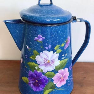 Peut inclure: Une cafetière en émail bleu avec un couvercle, une poignée et un bec verseur. La cafetière est décorée de pensées peintes à la main dans des tons de violet, de rose et de blanc, avec des feuilles vertes. La cafetière a une finition mouchetée.