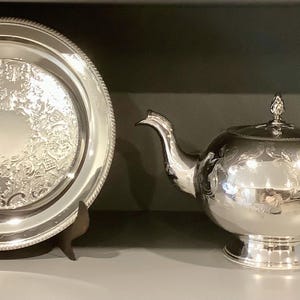 May include: A silver tea set comprising a teapot and a serving tray. The teapot features a curved spout and handle, with a decorative finial on the lid. The tray is round with an intricate pattern.